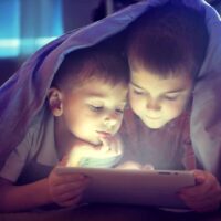 Two,Kids,Using,Tablet,Pc,Under,Blanket,At,Night.,Brothers